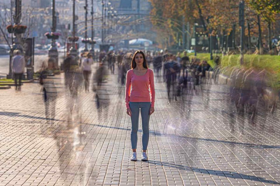 Femme en haut rose et jean bleu marchant seule sur un trottoir pavé dans une rue animée avec des passants flous.