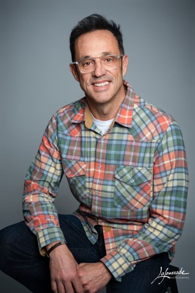 Homme souriant assis et décontracté, portant une chemise à carreaux colorée et des lunettes fines.