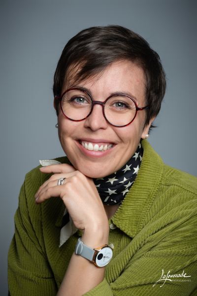 Femme souriante portant des lunettes rondes rouges, un pull vert et une écharpe noire à étoiles blanches.